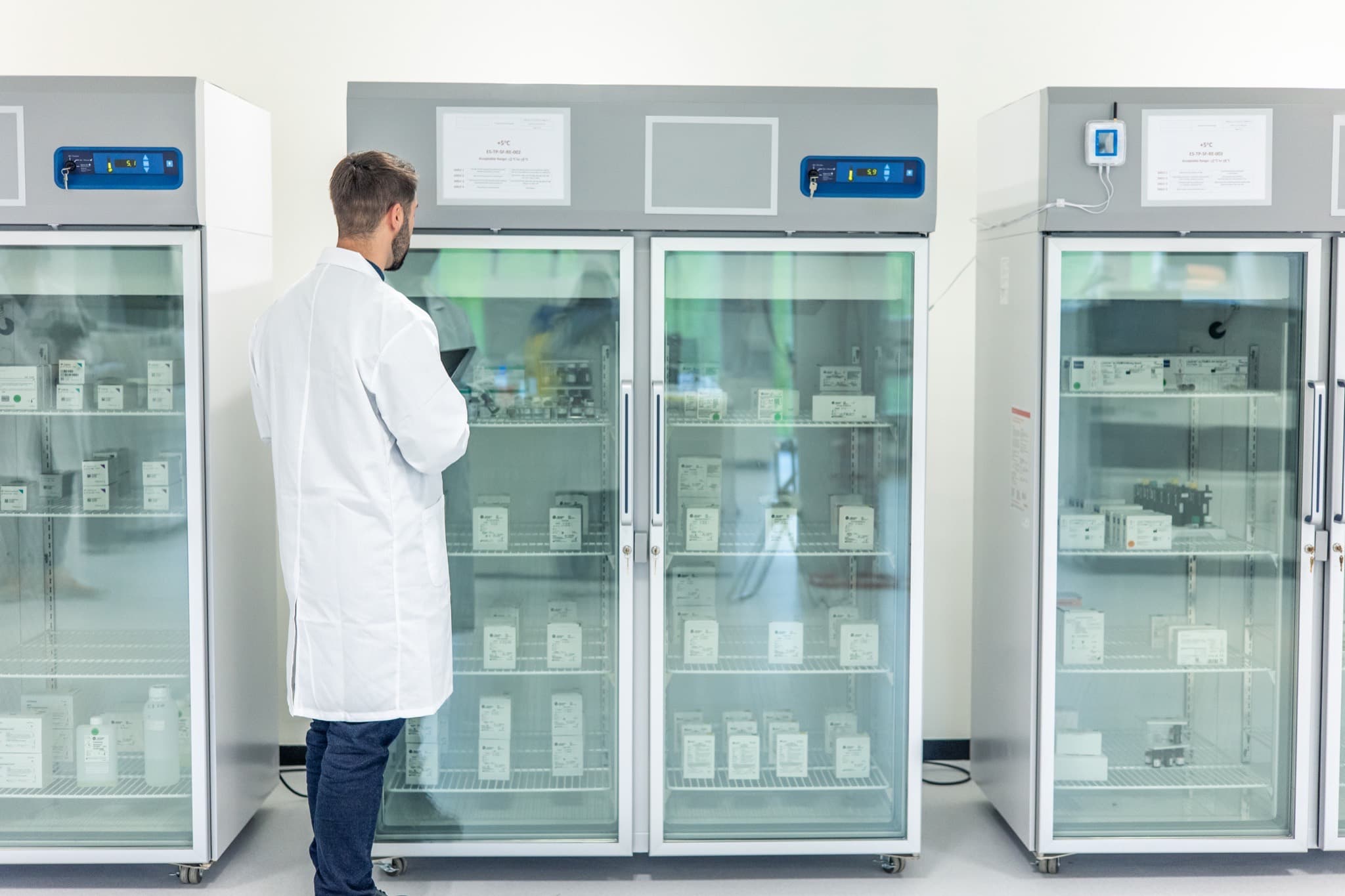 Three upright glass-door pharmaceutical refrigerators in a row with boxed stock on their shelves and digital temperature controllers above the doors, as a laboratory worker in a white coat checks the middle unit on a tablet