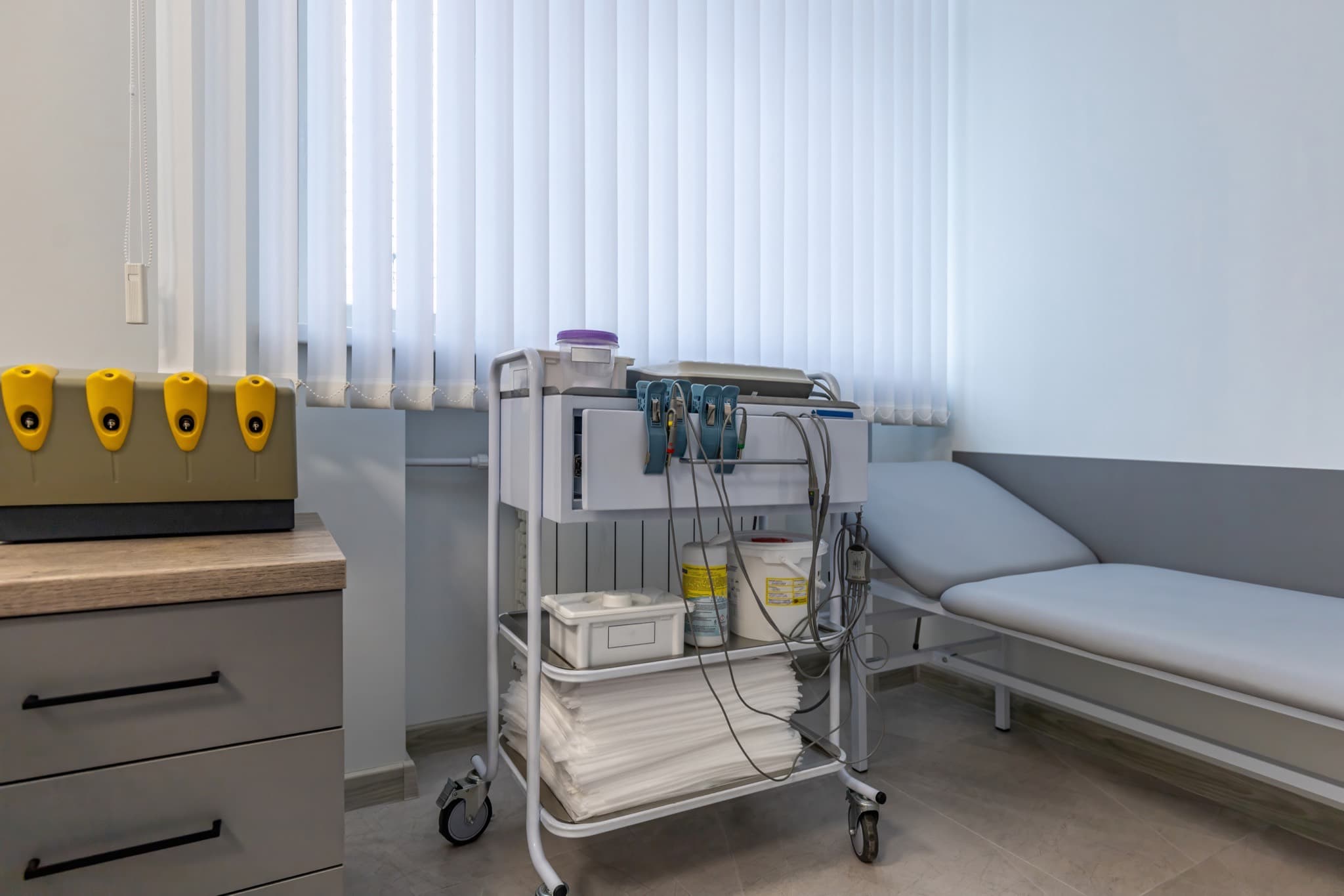 A white wheeled clinical trolley in a treatment room, with a top drawer, monitoring leads hanging from its front, and open shelves holding stacked paper couch covers, a storage tray and tubs, parked beside an examination couch and a grey drawer unit