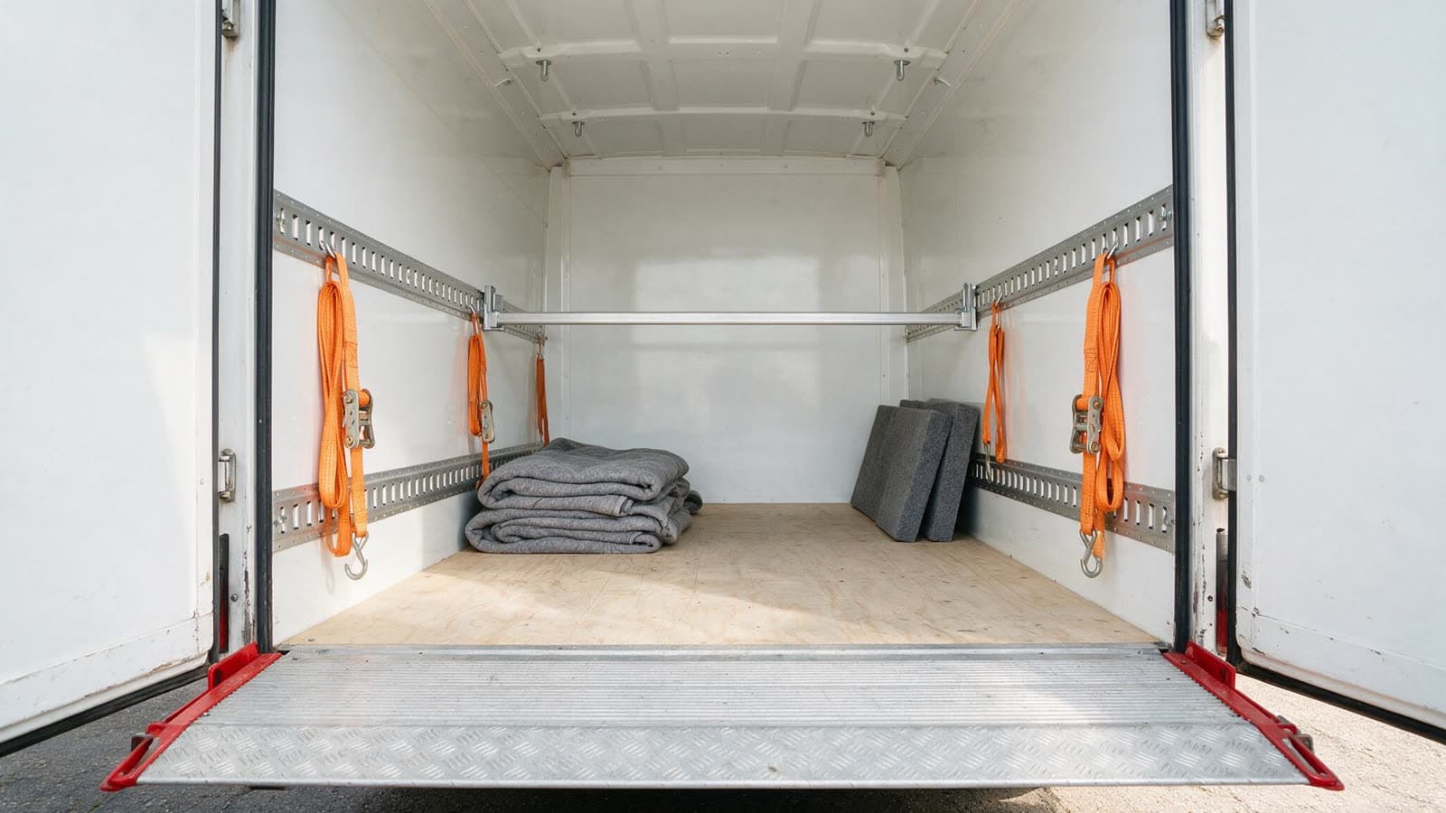 Interior of a box van fitted for medical equipment: aluminium lashing rails, coiled ratchet straps on hooks, a load bar braced across the body, folded moving blankets and foam blocks